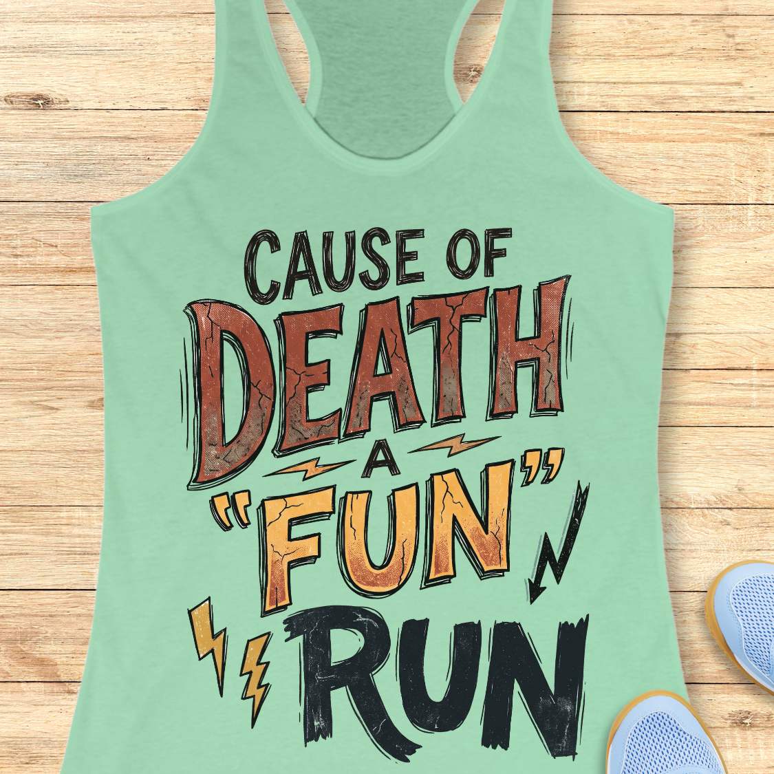 A mint green racerback tank top with a distressed graphic reading 'CAUSE OF DEATH A "FUN" RUN' in black, brown, and orange lettering, laid flat on a light wood plank background with partial sneakers visible at the edges.