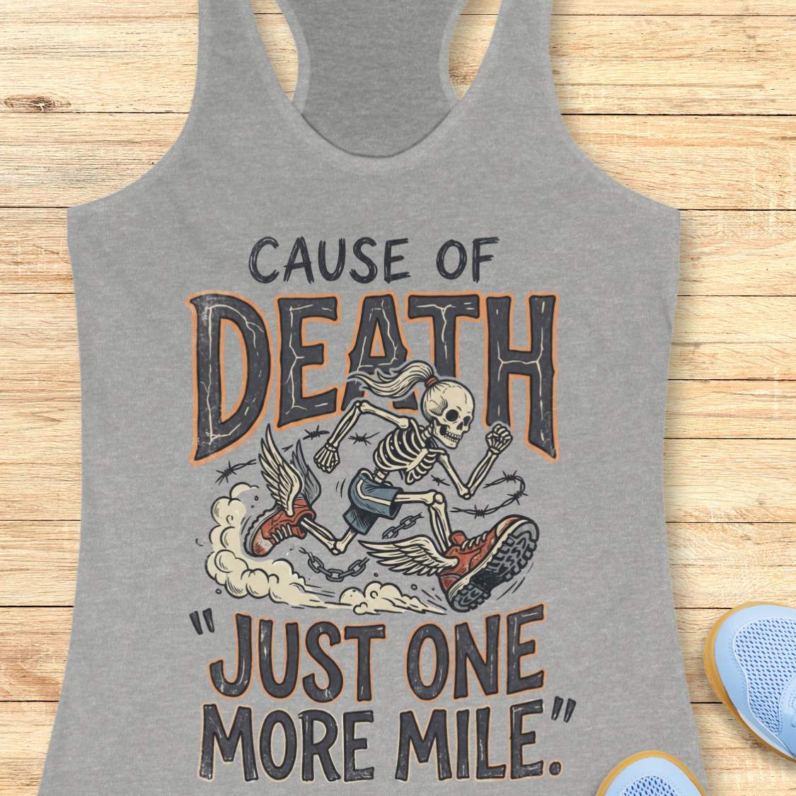 A heather gray racerback tank top with a graphic print of a skeleton running with a tire and the text 'CAUSE OF DEATH "JUST ONE MORE MILE".' The tank top is displayed flat on a light wood background with blue sneakers partially visible at the bottom corners.