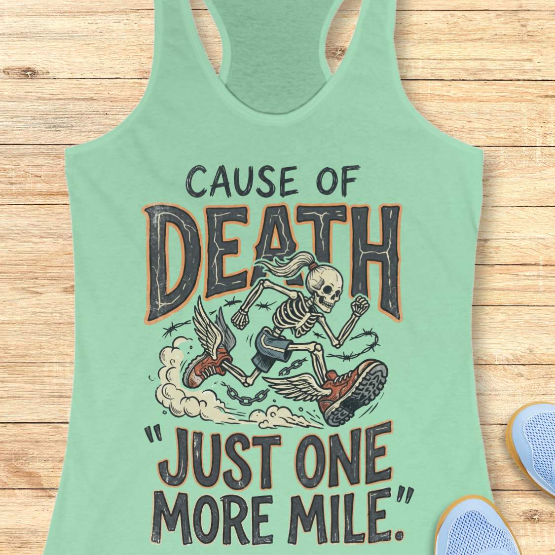 A mint green racerback tank top laid flat on a light wood background, featuring a graphic of a skeleton running with a boot and the text 'CAUSE OF DEATH "JUST ONE MORE MILE"'.