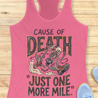 A pink racerback tank top laid flat on a light wood background, featuring a graphic of a skeleton running with a boot while being chased by a winged creature. The text reads 'CAUSE OF DEATH' and '"JUST ONE MORE MILE"'.