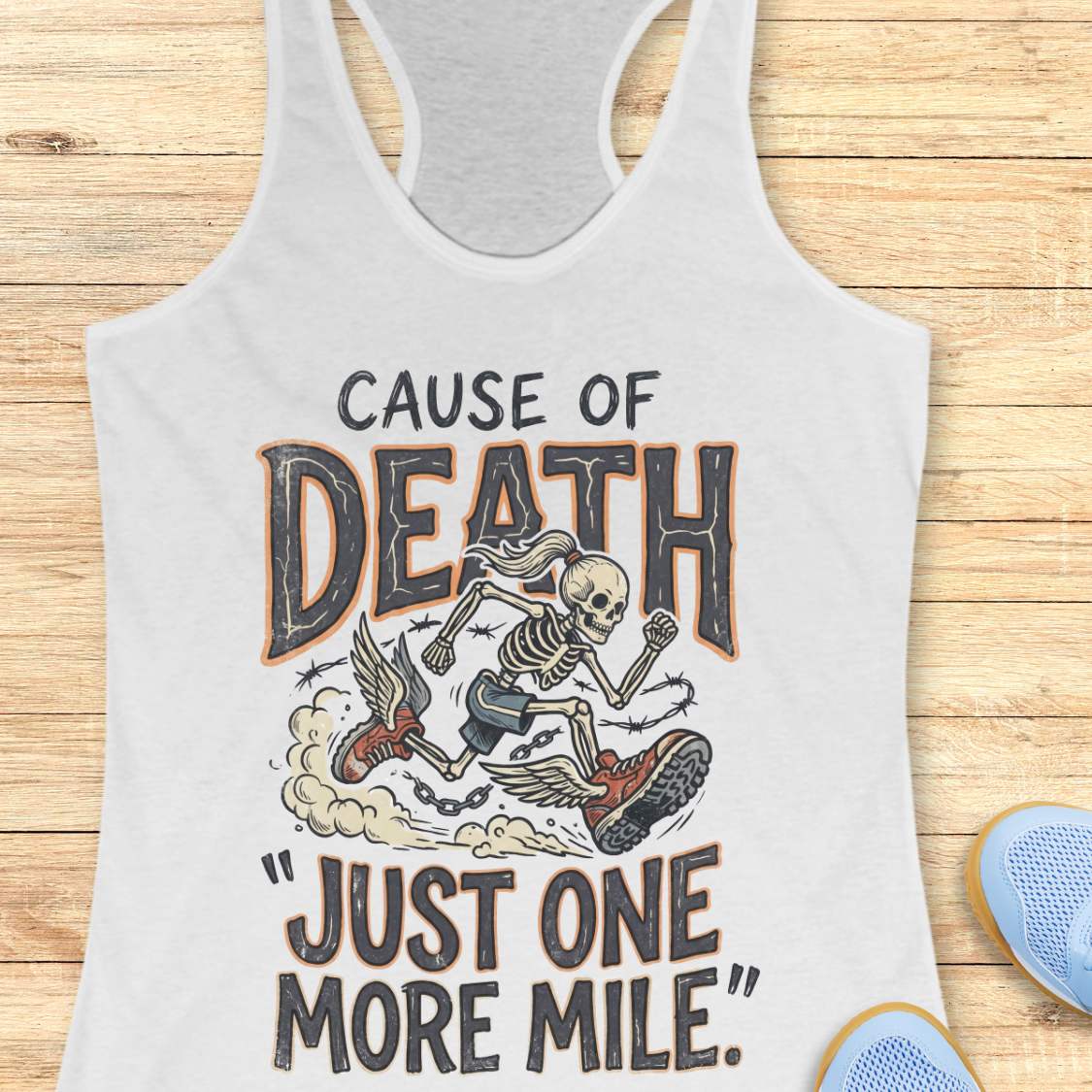 A white racerback tank top laid flat on a light wood background, featuring a graphic print of a skeleton running with a chain and the text 'CAUSE OF DEATH "JUST ONE MORE MILE"'. Partial blue shoes are visible at the bottom right as styling props.