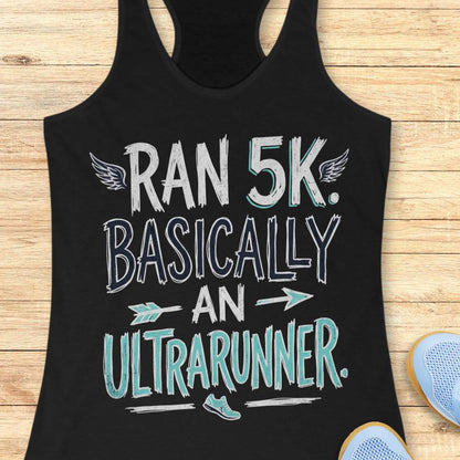 A black racerback tank top laid flat on a light wood background, featuring a graphic that reads 'RAN 5K. BASICALLY AN ULTRARUNNER.