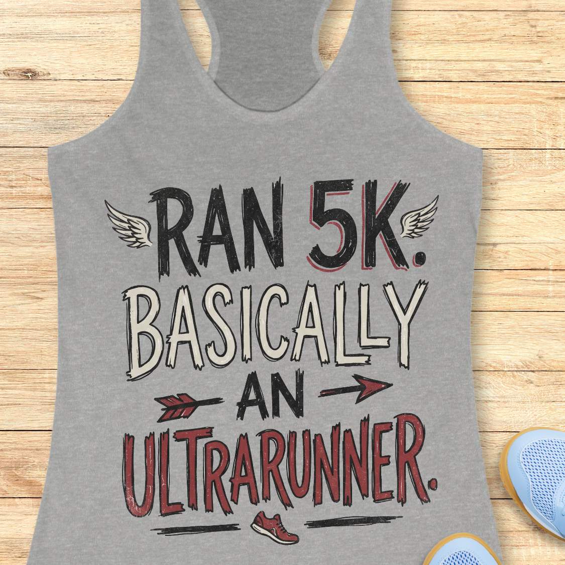 A heather gray racerback tank top laid flat on a light wood background, featuring the printed text 'RAN 5K. BASICALLY AN ULTRARUNNER.