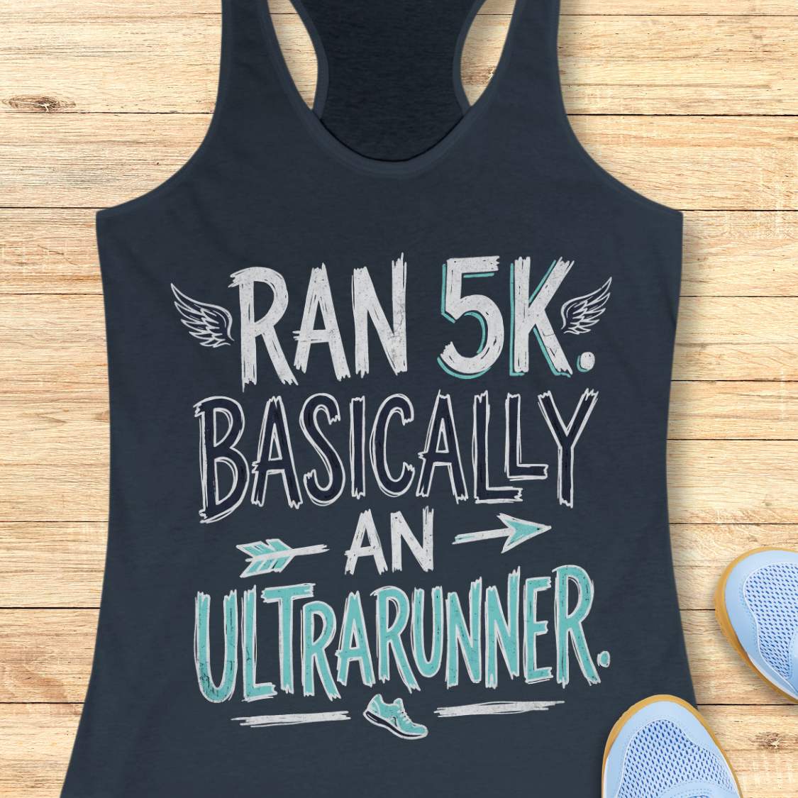 A indigo racerback tank top laid flat on a light wood background, printed with the phrase 'RAN 5K. BASICALLY AN ULTRARUNNER.