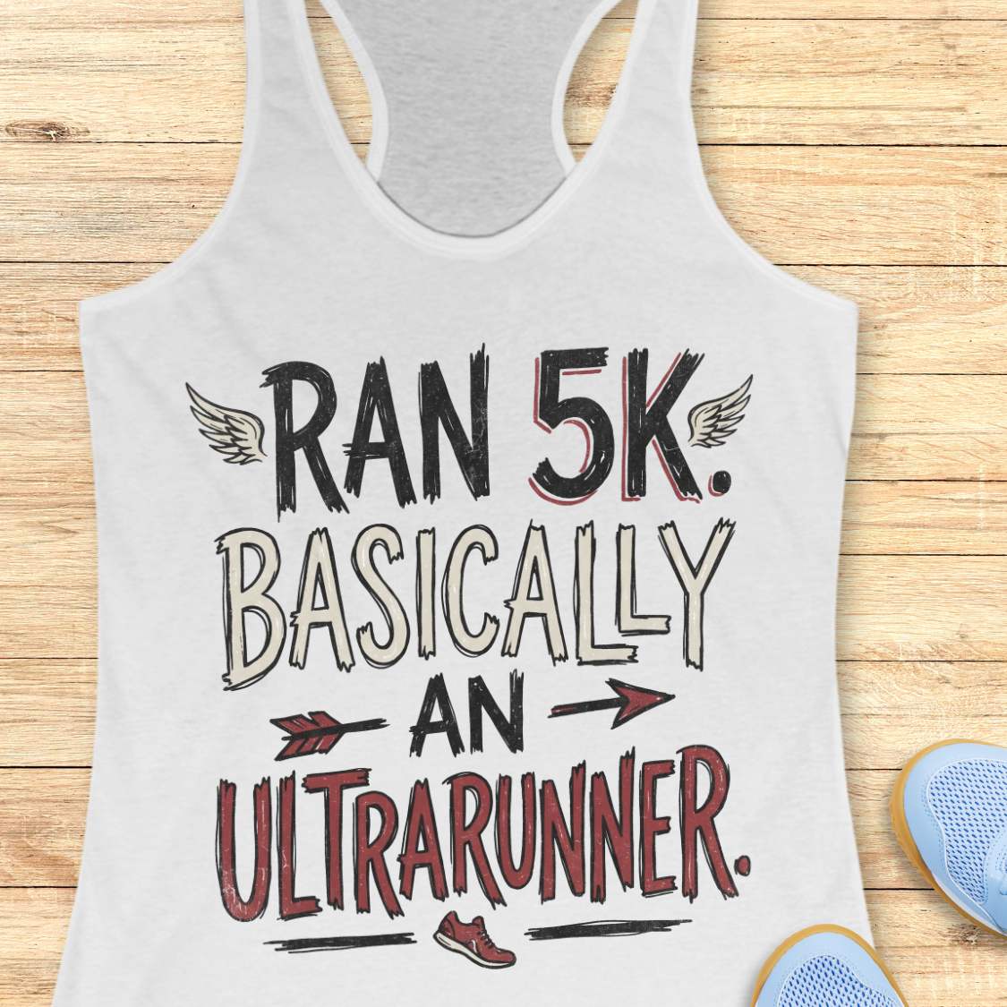 A white racerback tank top laid flat on a light wood plank background, featuring a graphic print that reads 'RAN 5K. BASICALLY AN ULTRARUNNER.