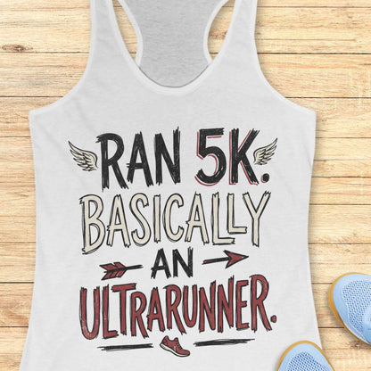 A white racerback tank top laid flat on a light wood plank background, featuring a graphic print that reads 'RAN 5K. BASICALLY AN ULTRARUNNER.
