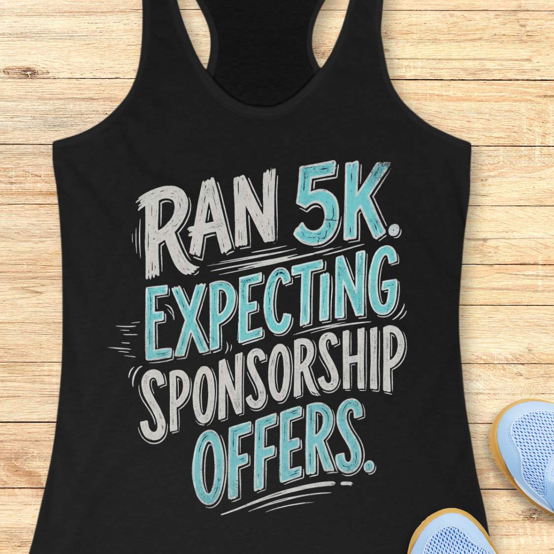 A black racerback tank top laid flat on a light wood background, featuring the text 'RAN 5K. EXPECTING SPONSORSHIP OFFERS.