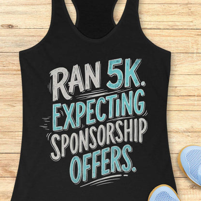 A black racerback tank top laid flat on a light wood background, featuring the text 'RAN 5K. EXPECTING SPONSORSHIP OFFERS.