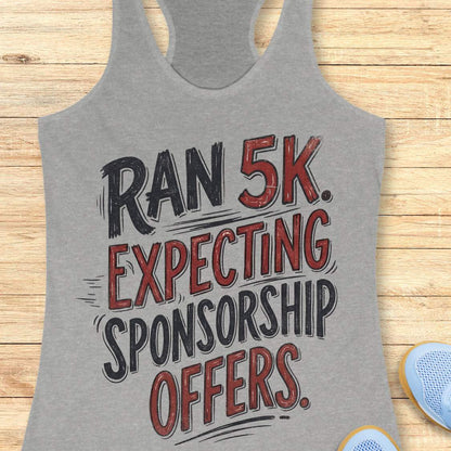 A heather gray racerback tank top laid flat on a light wood background, featuring the text 'RAN 5K. EXPECTING SPONSORSHIP OFFERS.