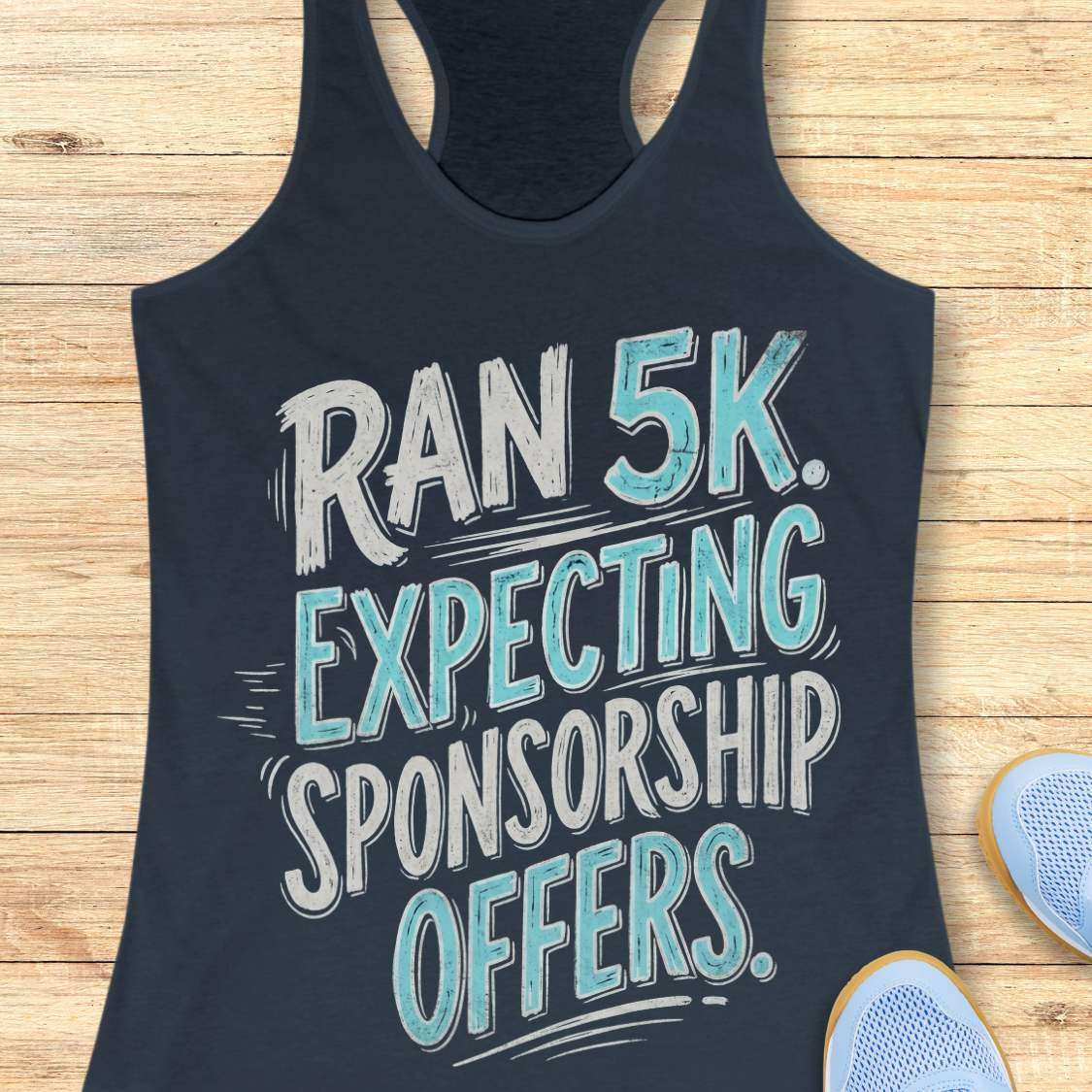 A indigo racerback tank top laid flat on a light wood background, printed with the phrase 'RAN 5K. EXPECTING SPONSORSHIP OFFERS.