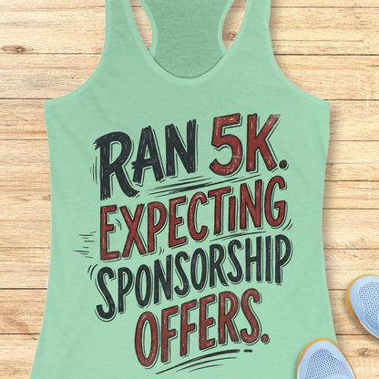 A mint green racerback tank top laid flat on a light wood plank background, featuring the text 'RAN 5K. EXPECTING SPONSORSHIP OFFERS.