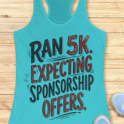 A turquoise racerback tank top laid flat on a light wood background, featuring the text 'RAN 5K. EXPECTING SPONSORSHIP OFFERS.