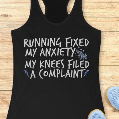 A black racerback tank top laid flat on a light wood surface, printed with the phrase 'RUNNING FIXED MY ANXIETY MY KNEES FILED A COMPLAINT' in black lettering, with small lightning bolt and wing graphics. Partial sneakers are visible at the bottom right as styling props.