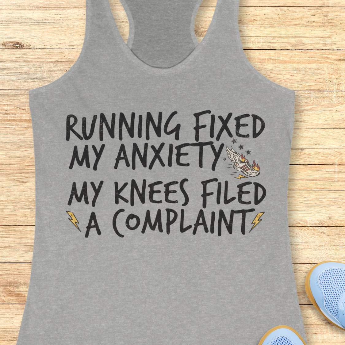 A heather gray racerback tank top with a humorous graphic printed on the front, laid flat on a light wood plank background. The graphic reads 'RUNNING FIXED MY ANXIETY MY KNEES FILED A COMPLAINT' in a black hand-drawn font, with small yellow lightning bolts and a small winged figure illustration.