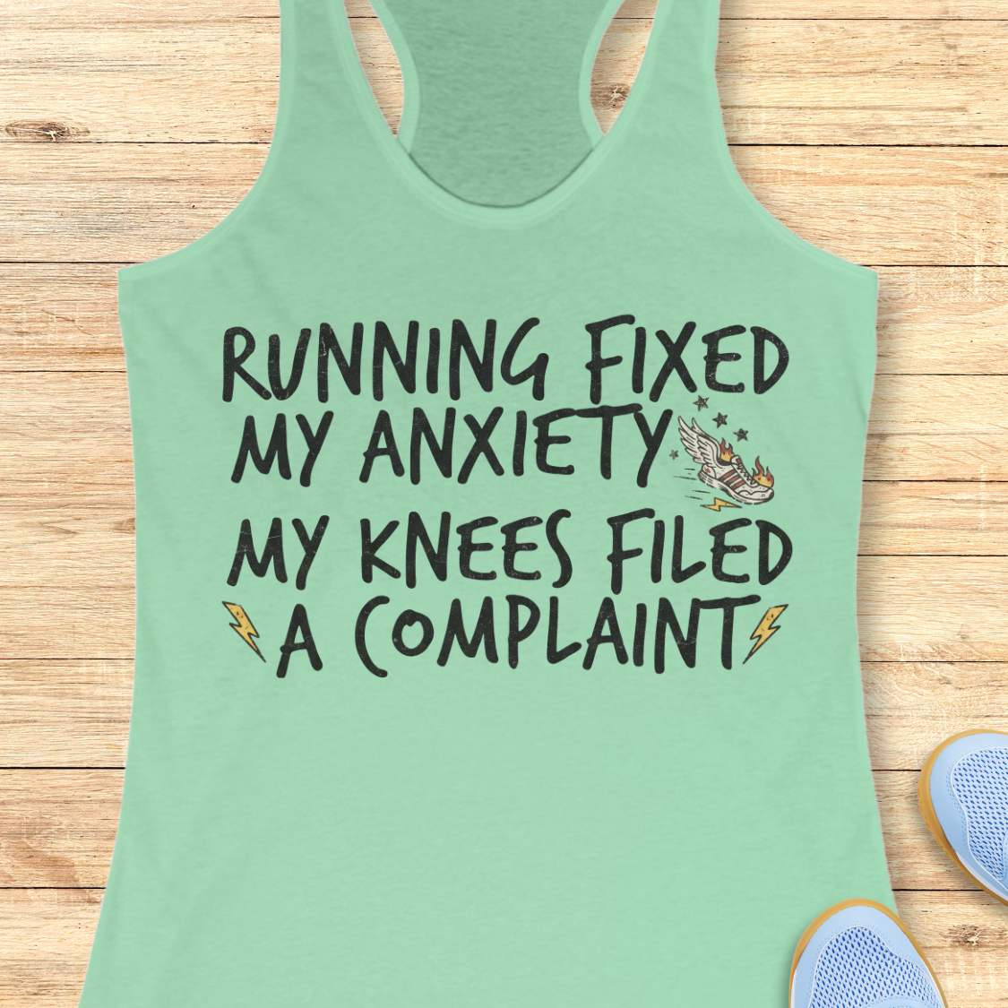 A mint green racerback tank top with a humorous graphic printed on the front, laid flat on a light wood background with partial blue sneakers visible at the bottom right.