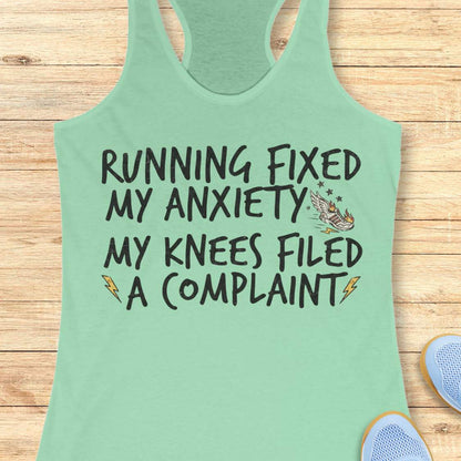 A mint green racerback tank top with a humorous graphic printed on the front, laid flat on a light wood background with partial blue sneakers visible at the bottom right.