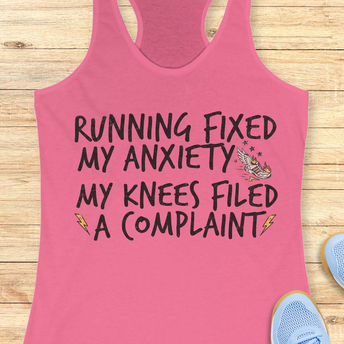 A pink racerback tank top laid flat on a light wood background, with black text reading “RUNNING FIXED MY ANXIETY MY KNEES FILED A COMPLAINT” and small graphic accents. Partial blue sneakers are visible at the bottom right as styling props.