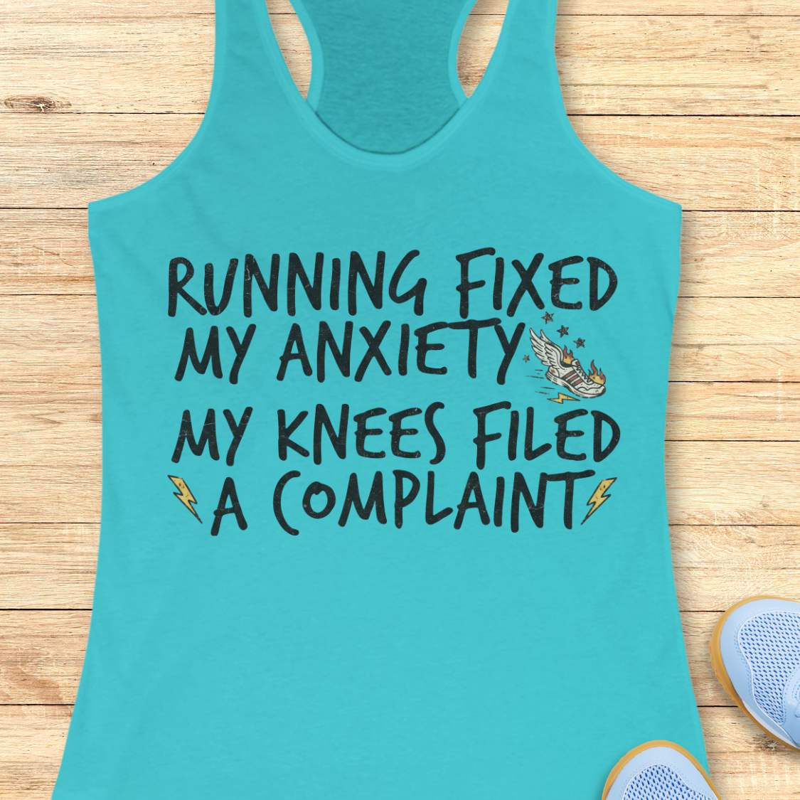 A turquoise racerback tank top laid flat on a light wood surface, printed with the phrase 'RUNNING FIXED MY ANXIETY MY KNEES FILED A COMPLAINT' in black lettering, with small lightning bolt and wing graphics. Partial sneakers are visible at the bottom right as styling props.