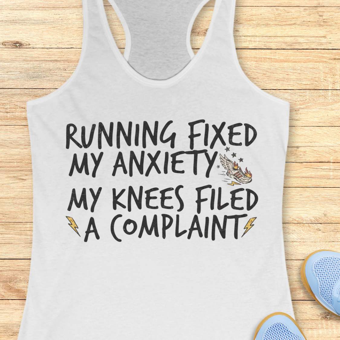 A white racerback tank top laid flat on a light wood plank background, featuring the printed text 'RUNNING FIXED MY ANXIETY MY KNEES FILED A COMPLAINT' in black, with small lightning bolt and eagle graphics. Partial blue shoes are visible at the bottom right as styling props.