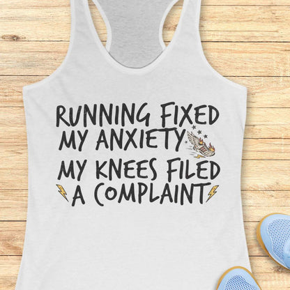 A white racerback tank top laid flat on a light wood plank background, featuring the printed text 'RUNNING FIXED MY ANXIETY MY KNEES FILED A COMPLAINT' in black, with small lightning bolt and eagle graphics. Partial blue shoes are visible at the bottom right as styling props.