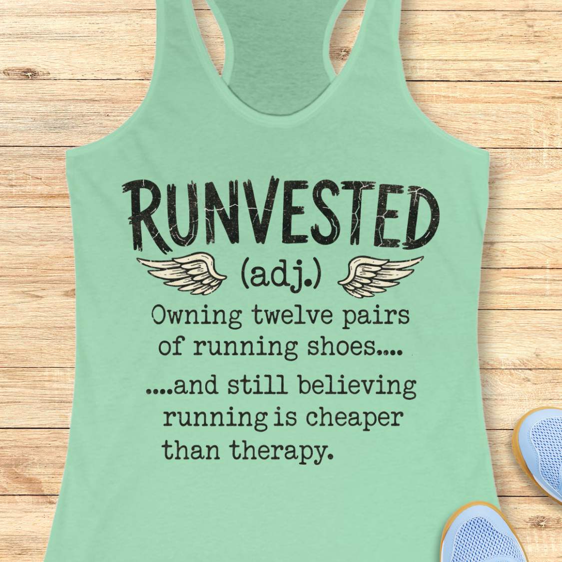 A mint green racerback tank top laid flat on a light wood background, featuring a humorous printed definition of 'RUNVESTED (adj.)' with wings and text about owning twelve pairs of running shoes.