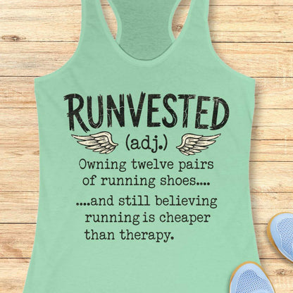 A mint green racerback tank top laid flat on a light wood background, featuring a humorous printed definition of 'RUNVESTED (adj.)' with wings and text about owning twelve pairs of running shoes.