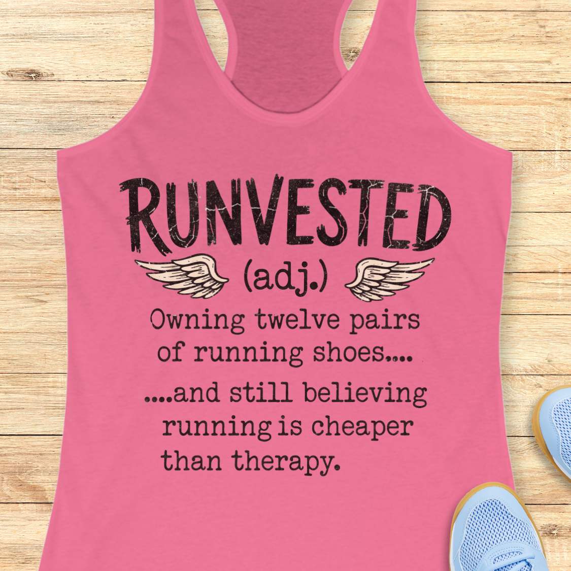 A pink racerback tank top laid flat on a light wood background, featuring black printed text that reads 'RUNVESTED (adj.)' with wing graphics and a humorous running quote.