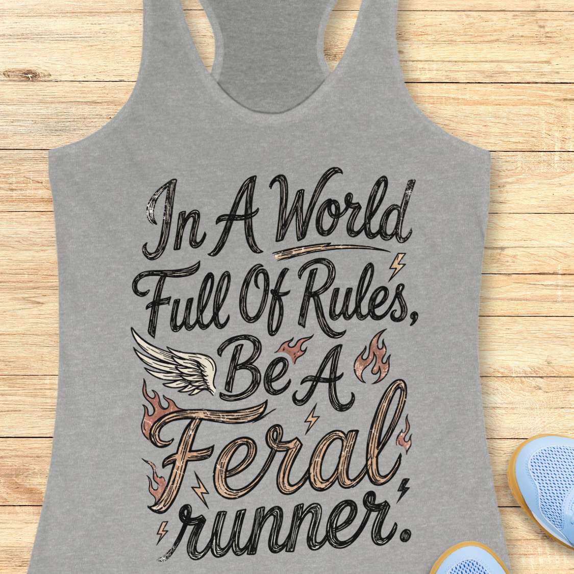 A grey racerback tank top laid flat on a light wood surface, featuring a distressed white and blue graphic that reads, 'In A World Full Of Rules, Be A Feral runner.' with a wing and flame motifs.