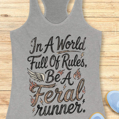 A grey racerback tank top laid flat on a light wood surface, featuring a distressed white and blue graphic that reads, 'In A World Full Of Rules, Be A Feral runner.' with a wing and flame motifs.