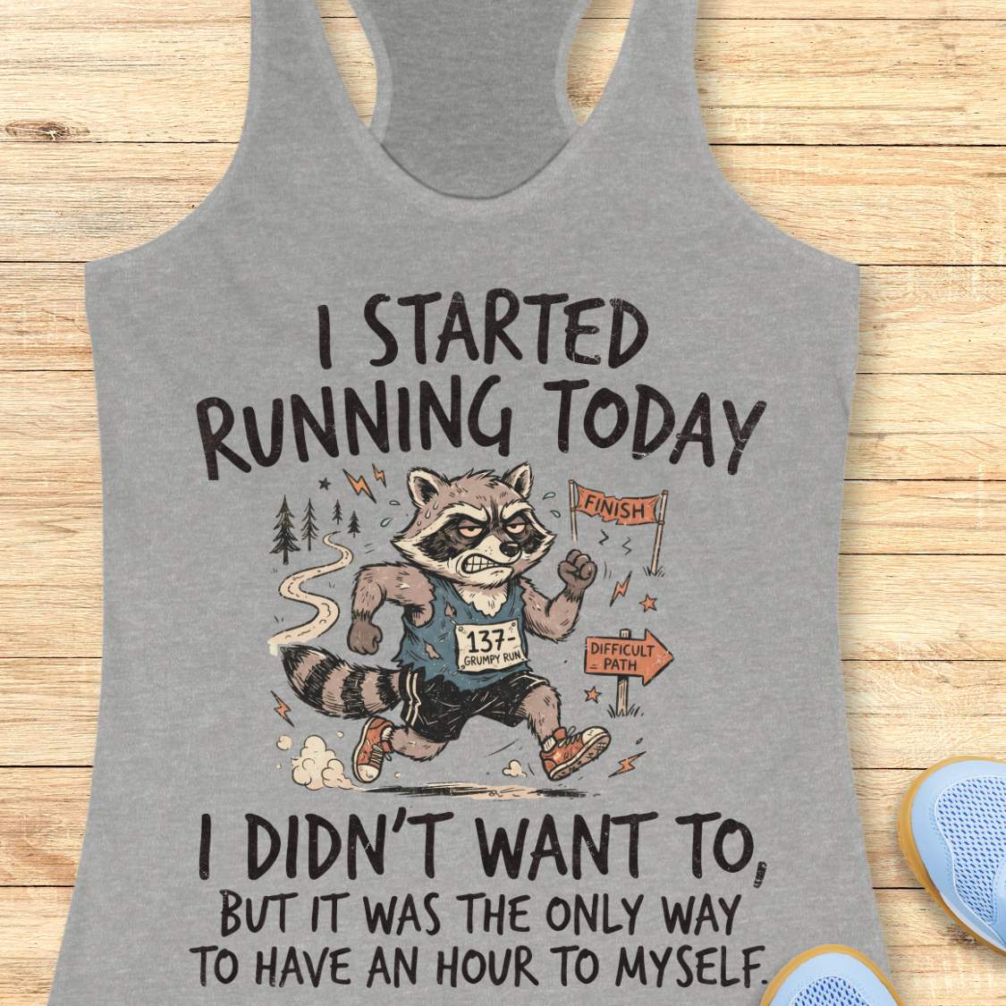 A heather gray racerback tank top laid flat on a light wood background, featuring a humorous graphic of a raccoon running with the text 'I STARTED RUNNING TODAY I DIDN'T WANT TO, BUT IT WAS THE ONLY WAY TO HAVE AN HOUR TO MYSELF'.