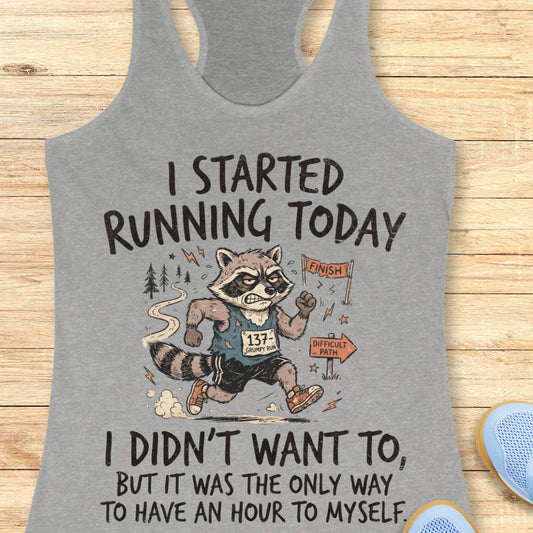 A heather gray racerback tank top laid flat on a light wood background, featuring a humorous graphic of a raccoon running with the text 'I STARTED RUNNING TODAY I DIDN'T WANT TO, BUT IT WAS THE ONLY WAY TO HAVE AN HOUR TO MYSELF'.