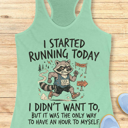 A mint green racerback tank top with a humorous raccoon running graphic and text, displayed flat on a light wood background with partial blue sneakers visible at the edges.