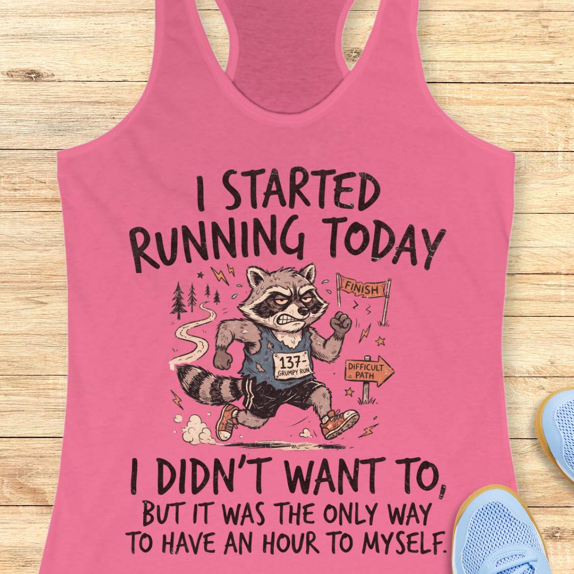 A pink racerback tank top with a humorous raccoon running graphic and text, displayed flat on a light wood background with partial blue sneakers visible at the edges.