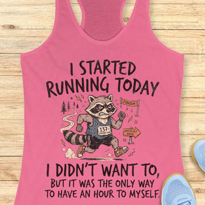 A pink racerback tank top with a humorous raccoon running graphic and text, displayed flat on a light wood background with partial blue sneakers visible at the edges.