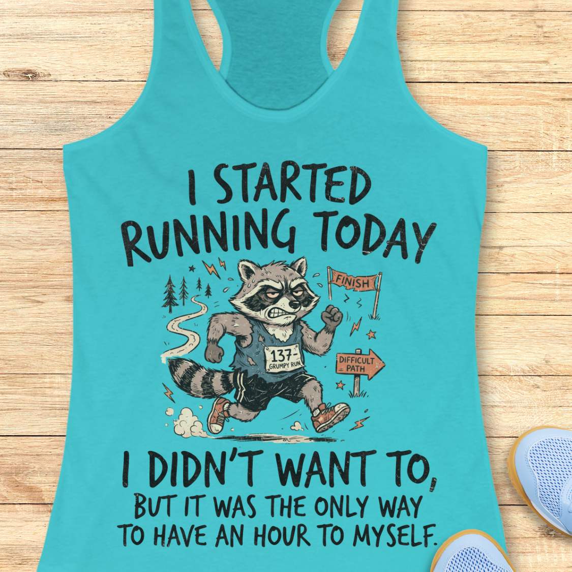 A turquoise racerback tank top laid flat on a light wood surface, featuring a humorous running graphic of a raccoon wearing a bib and the text 'I STARTED RUNNING TODAY I DIDN'T WANT TO, BUT IT WAS THE ONLY WAY TO HAVE AN HOUR TO MYSELF.' A partial blue shoe is visible at the bottom right.