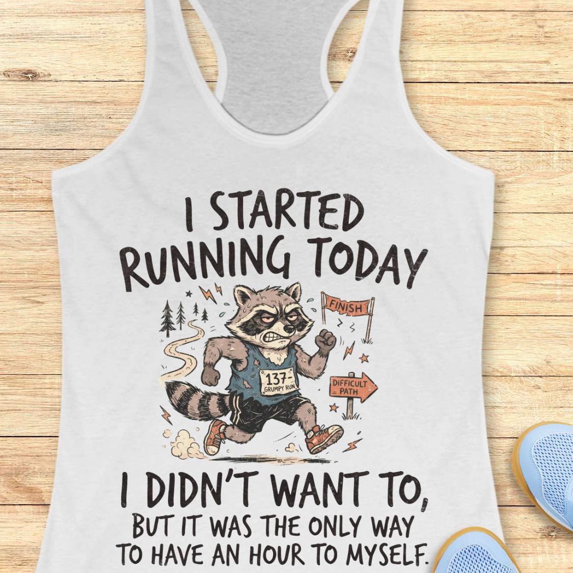 A white racerback tank top laid flat on a light wood background, featuring a humorous graphic of a raccoon running a race with the text 'I STARTED RUNNING TODAY I DIDN'T WANT TO, BUT IT WAS THE ONLY WAY TO HAVE AN HOUR TO MYSELF.' Partial blue shoes are visible at the bottom right edge.