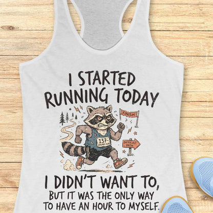A white racerback tank top laid flat on a light wood background, featuring a humorous graphic of a raccoon running a race with the text 'I STARTED RUNNING TODAY I DIDN'T WANT TO, BUT IT WAS THE ONLY WAY TO HAVE AN HOUR TO MYSELF.' Partial blue shoes are visible at the bottom right edge.