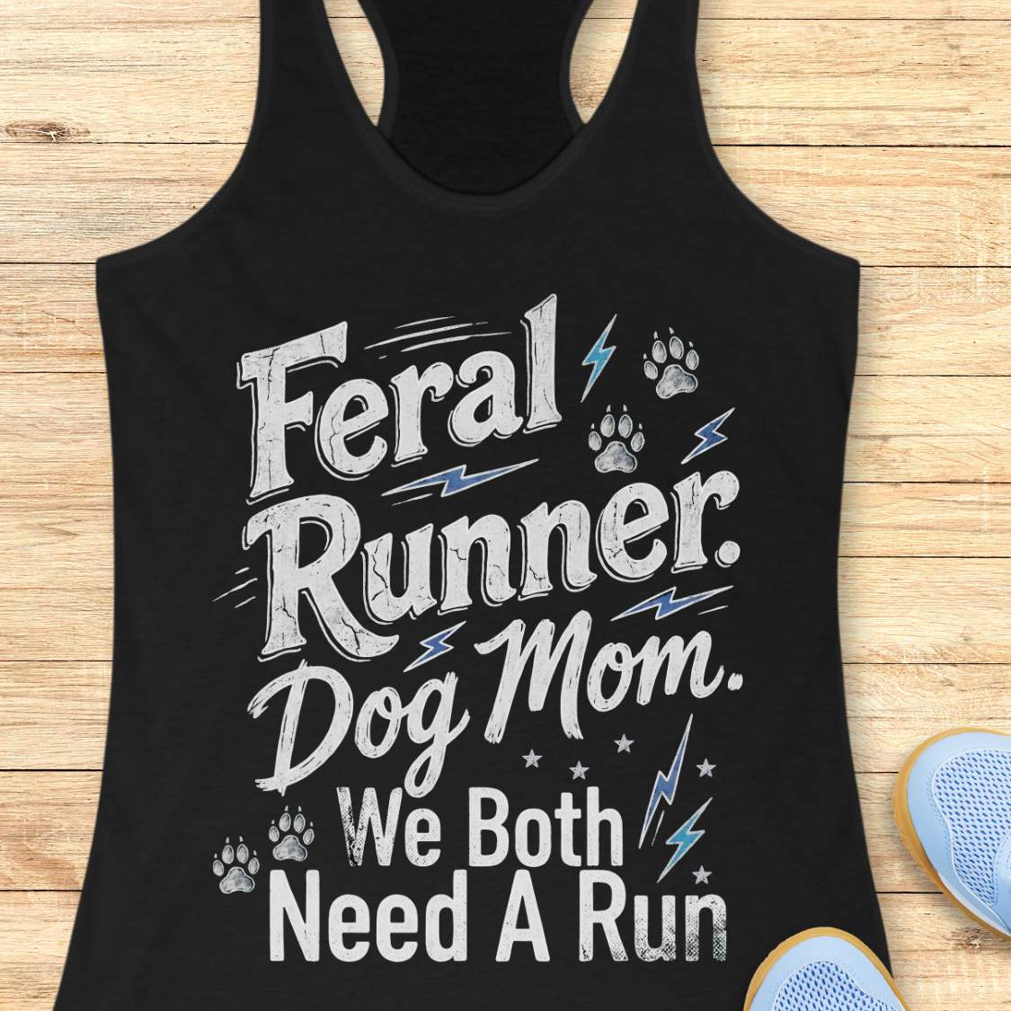 A black racerback tank top laid flat on a light wood background, printed with the phrase 'Feral Runner Dog Mom We Both Need A Run' in distressed white lettering with small paw prints, lightning bolts, and stars. Part of a blue shoe is visible at the bottom right.