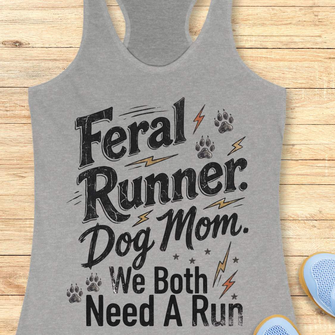 A heather gray racerback tank top laid flat on a light wood background, featuring the printed phrase 'Feral Runner. Dog Mom.