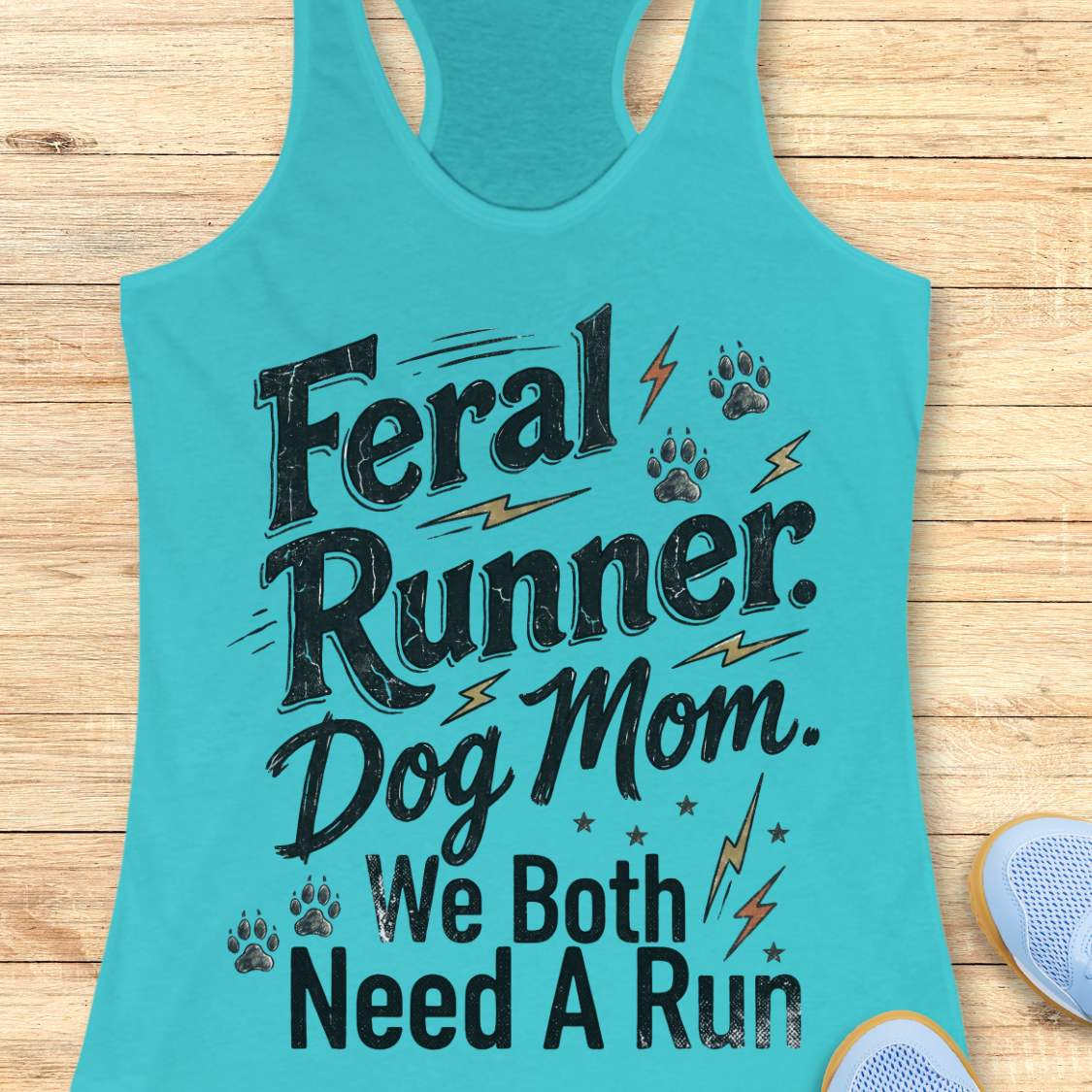 A turquoise racerback tank top laid flat on a light wood surface, printed with the phrase 'Feral Runner. Dog Mom.