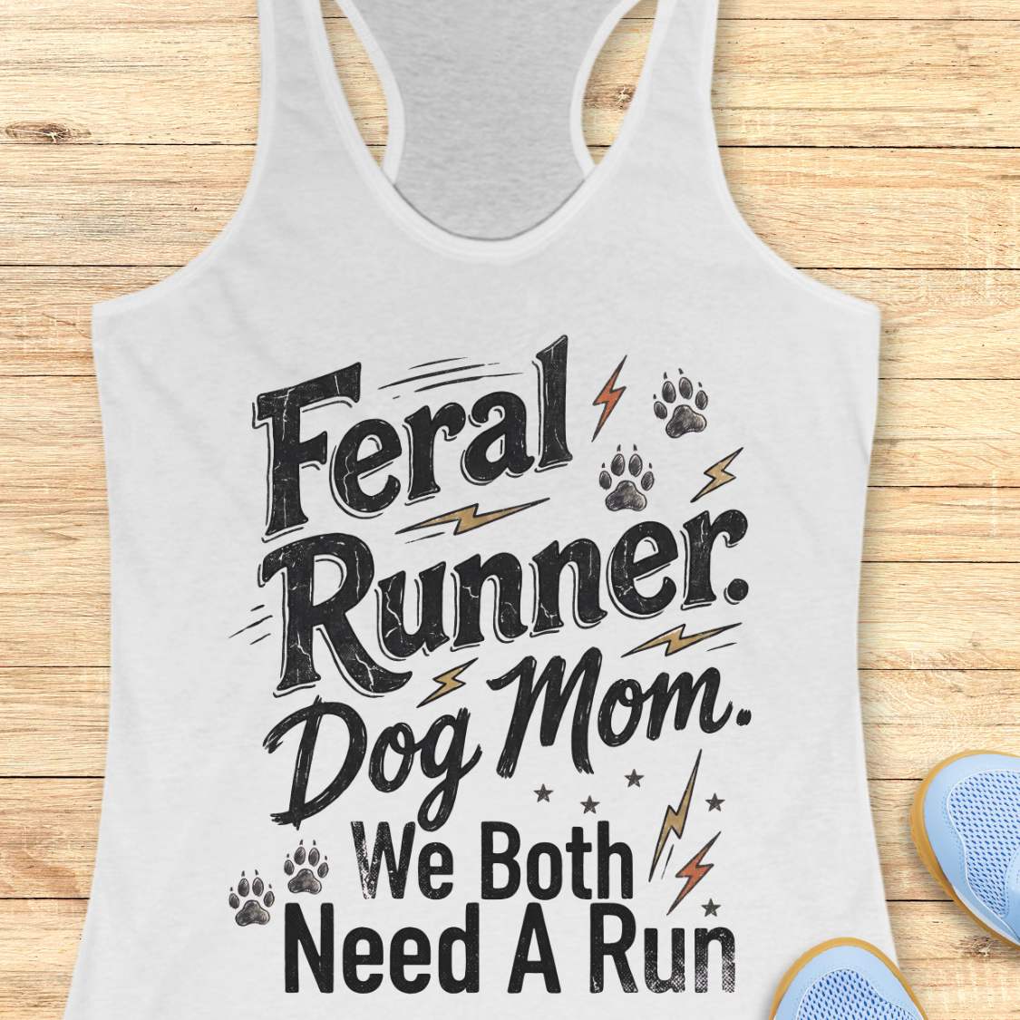 A white racerback tank top laid flat on a light wood background, featuring the printed phrase 'Feral Runner. Dog Mom.