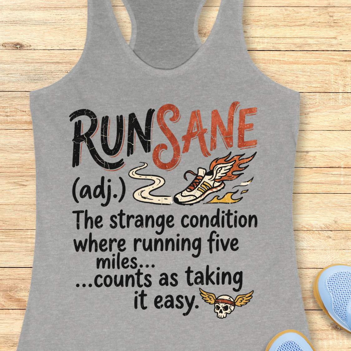 A heather gray racerback tank top laid flat on a light wood background, featuring a humorous graphic print that says 'RUN SANE (adj.) The strange condition where running five miles.
