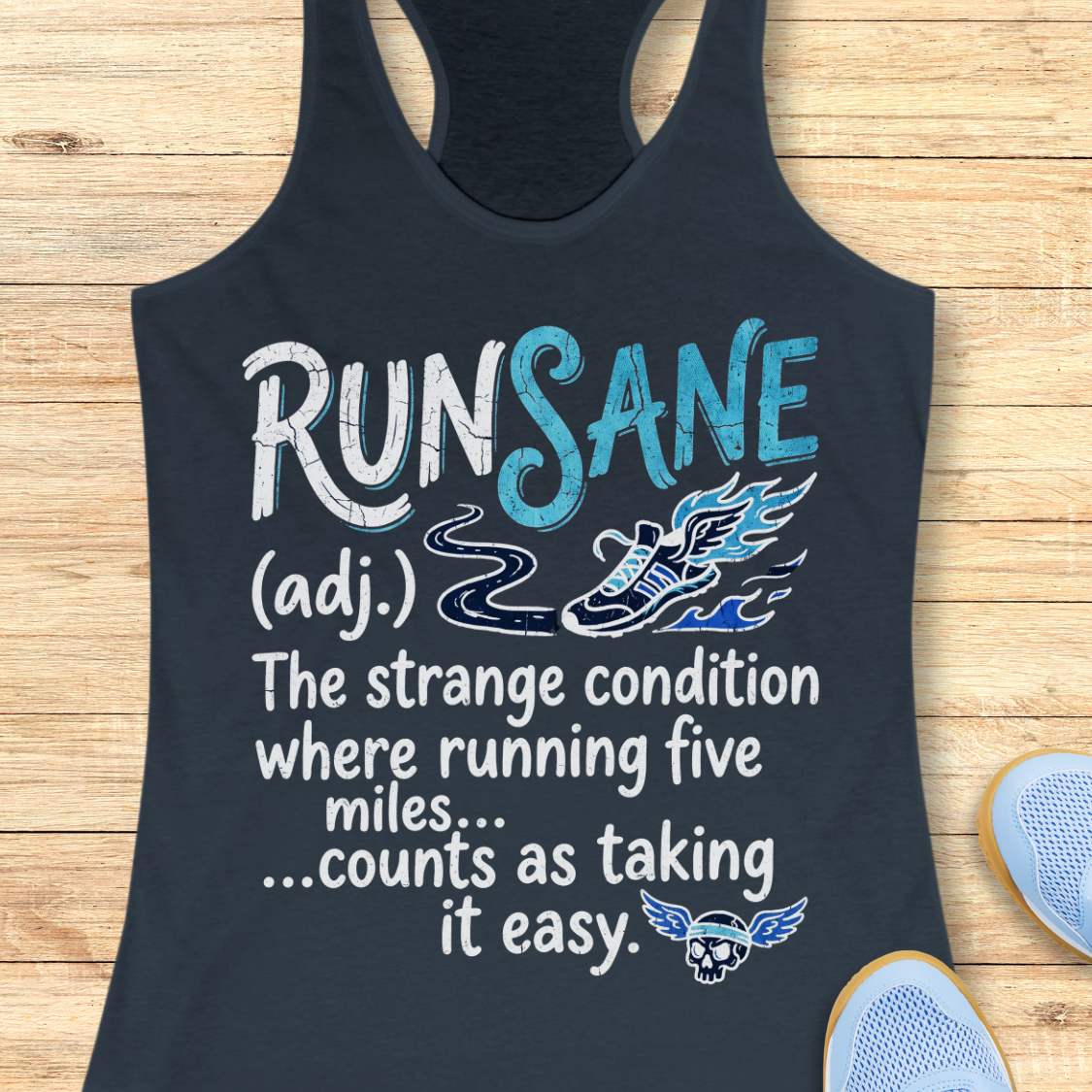 A indigo racerback tank top laid flat on a light wood background, featuring a white and blue graphic print that reads 'RUN SANE (adj.) The strange condition where running five miles .