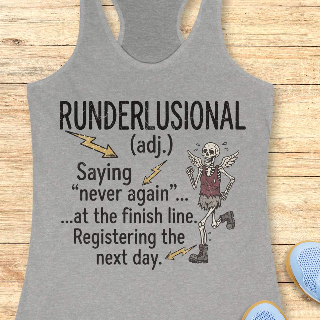 A heather gray racerback tank top laid flat on a light wood background, featuring a humorous dictionary-style definition print that reads 'RUNDERLUSIONAL (adj.) Saying "never again" .