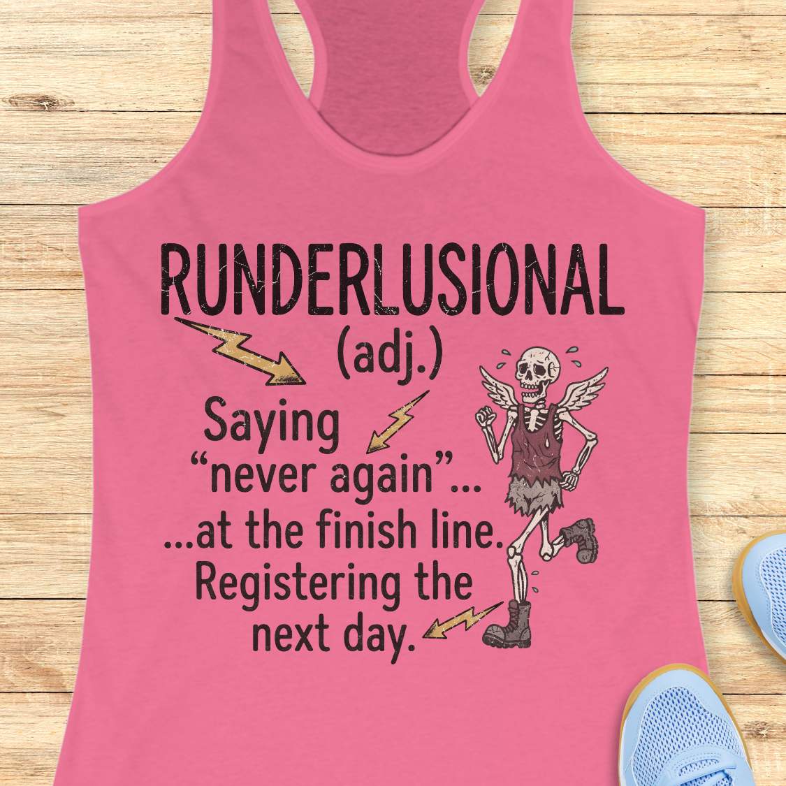 A pink racerback tank top laid flat on a light wood background, featuring a humorous graphic with the text 'RUNDERLUSIONAL (adj.) Saying "never again" .