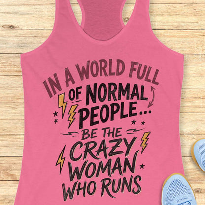 A pink racerback tank top with a motivational graphic printed on the front, laid flat on a light wood background with partial sneakers visible at the edges.