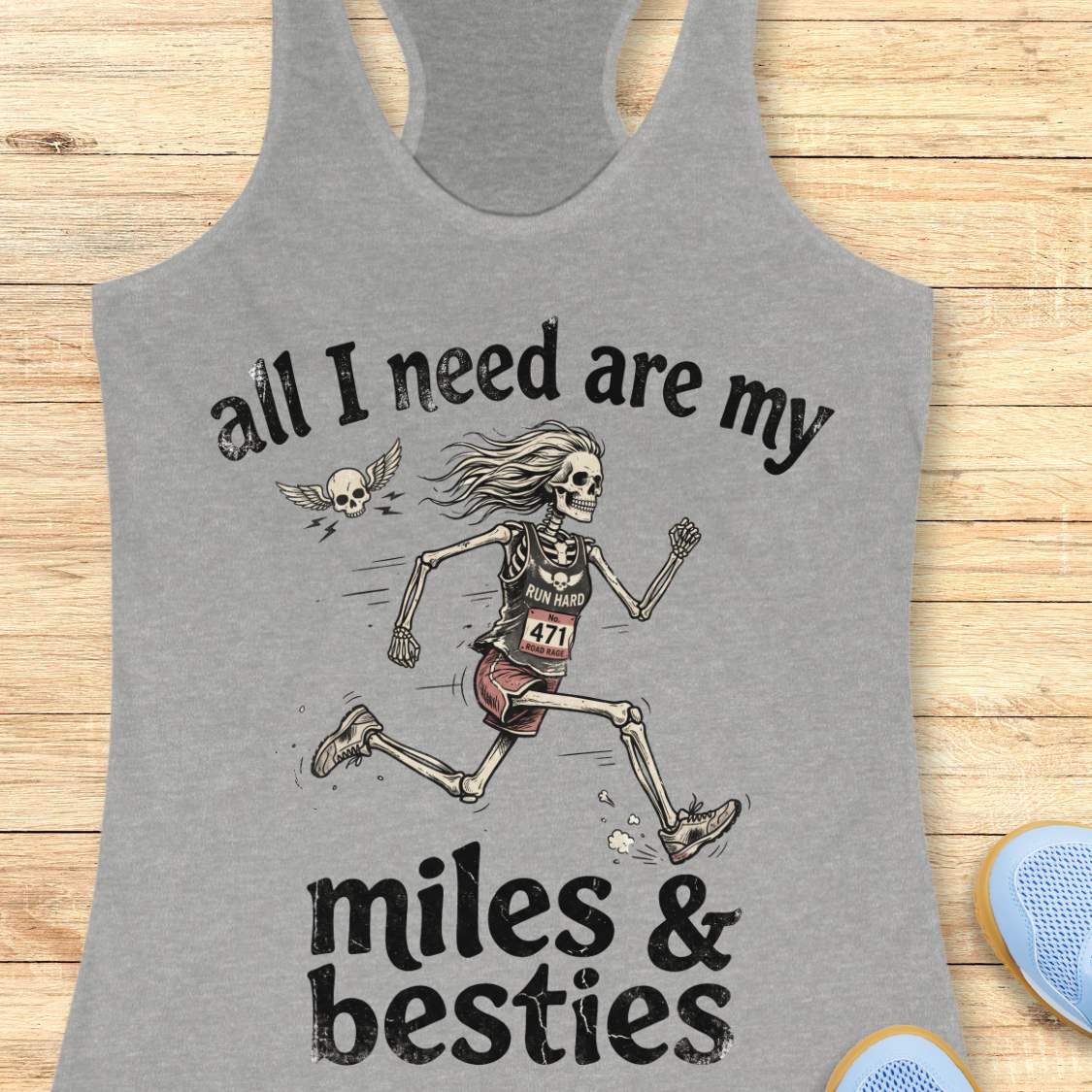 A heather grey racerback tank top laid flat on a light wood background, featuring a colorful graphic that reads 'all I need are my miles & besties' with a running female skeleton. Part of a blue shoe is visible at the bottom right.