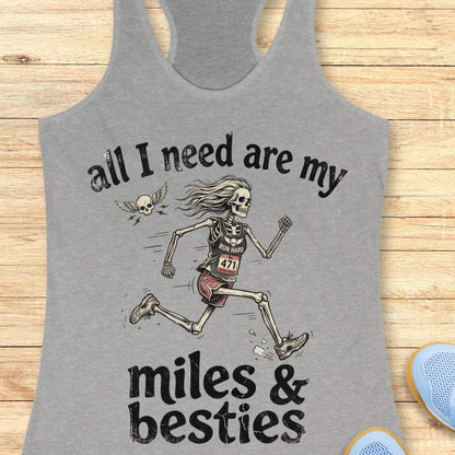 A heather grey racerback tank top laid flat on a light wood background, featuring a colorful graphic that reads 'all I need are my miles & besties' with a running female skeleton. Part of a blue shoe is visible at the bottom right.