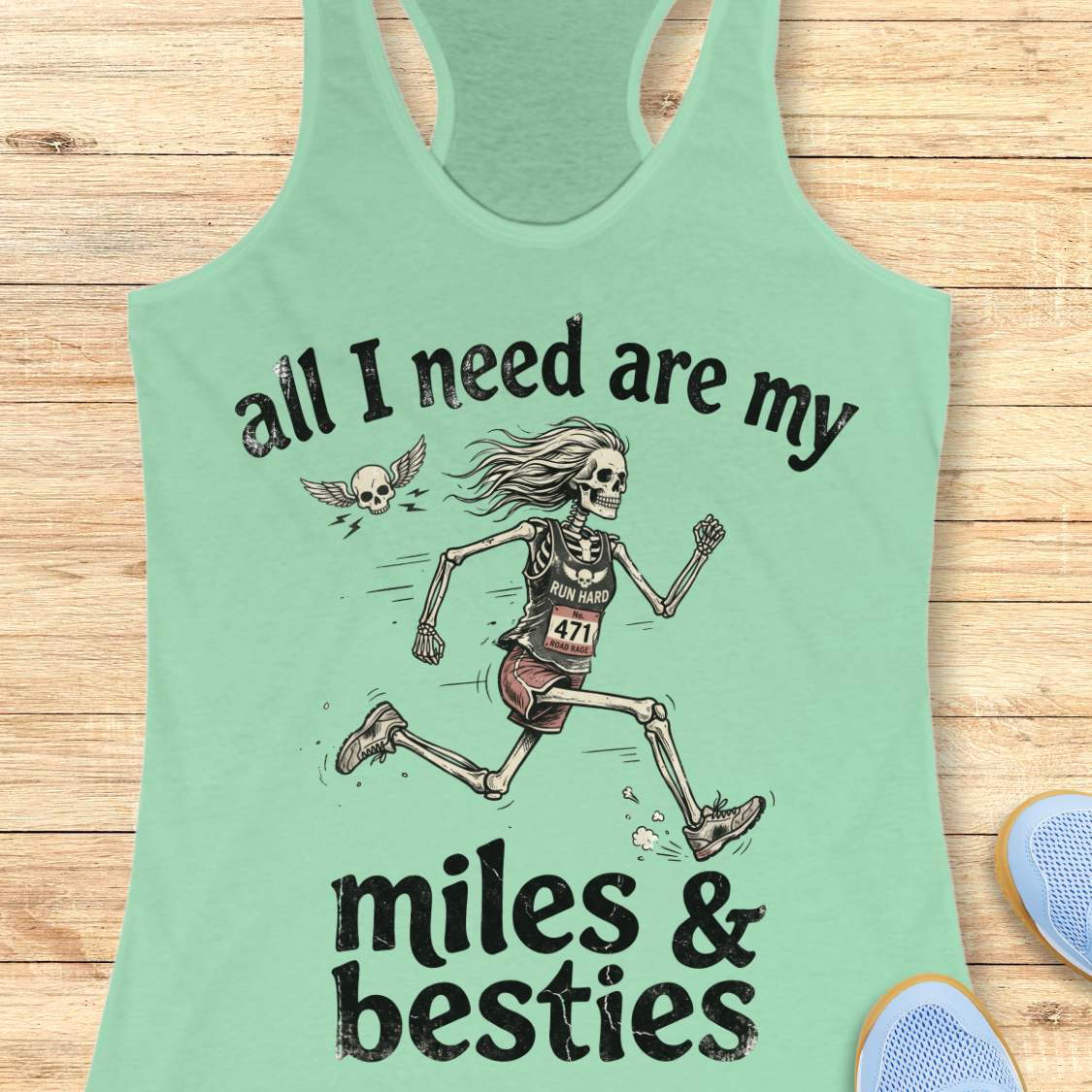 A mint racerback tank top laid flat on a light wood background, featuring a colorful graphic that reads 'all I need are my miles & besties' with a running female skeleton. Part of a blue shoe is visible at the bottom right.