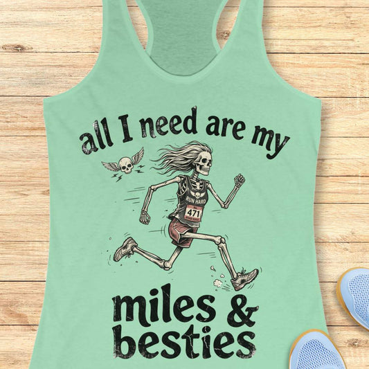 A mint racerback tank top laid flat on a light wood background, featuring a colorful graphic that reads 'all I need are my miles & besties' with a running female skeleton. Part of a blue shoe is visible at the bottom right.